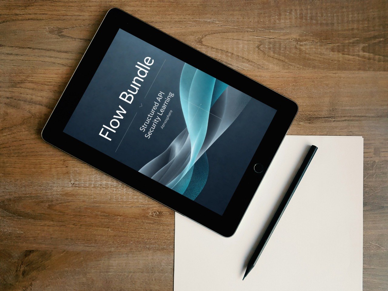 A tablet lying on a wooden desk next to a blank sheet of paper and a stylus, with the screen displaying the text 'Flow Bundle' and a dark abstract graphic.
